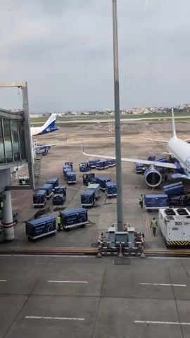 The video shows an airport tarmac scene with multiple airplanes and baggage carts. In the foreground, a jet bridge extends from a terminal building. Several blue baggage carts are arranged in a U-shape in front of a white and blue airplane. Two ground crew members in high-visibility vests are visible near the baggage carts and the aircraft. Another airplane, also blue and white, is parked further in the background. The tarmac appears to be paved with asphalt. The sky is overcast with a few clouds visible.