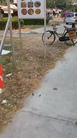 A black bicycle with a yellow box on the back labeled "SOL SEPATU" is parked next to a sidewalk. The bicycle has a basket on the front and a light-colored seat. In the background, there are buildings, trees, and a silver car driving down the road. The ground is a mix of dirt and pavement.