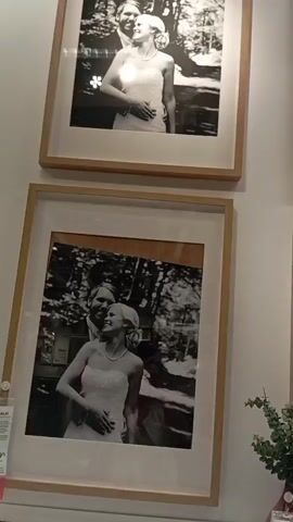 A video shows a wall decorated with several framed black and white photographs of a wedding couple. The photographs are arranged in a grid, with two rows of two pictures each visible. Each picture features a smiling bride in a white wedding dress and a groom embracing her. The frames are light wood. In the lower right corner, a small bouquet of green leaves and a pink flower is visible. The wall is white. The video pans slightly across the photographs.