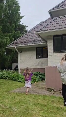 This media file depicts a backyard scene in Harkujärvi, Estonia. The primary subjects are a young girl with long brown hair, wearing a purple top and a patterned skirt, and a woman with blonde hair, seen from behind, wearing a striped shirt and black pants. The girl is walking away from the camera towards a brick house with a brown tiled roof and white trim. She appears to be barefoot. The woman is standing on the right side of the frame, facing the house.

The setting is a grassy yard with a paved walkway leading towards the house. Lush green bushes and trees surround the area, with a particularly large birch tree visible in the background. The weather appears overcast, suggesting it might be a cloudy day. The overall mood is casual and domestic, possibly indicating a family outing or a moment of play. There are no visible text elements in the video. The scene is captured during daylight hours.