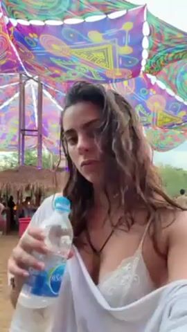The video shows a woman at an outdoor event, likely a festival or party, drinking from a plastic water bottle. She has long, wavy, dark hair and is wearing a white cover-up over a lace top. The background is filled with people, music, and colorful decorations, including a large canopy overhead with psychedelic patterns. The ground appears to be dirt or sand. The woman takes a drink of water, then looks at the camera.