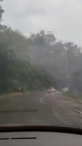 This media file captures a drive through a wooded, rural area in Briukhovychi, Ukraine, during a rainstorm. The perspective is from inside a vehicle, with the windshield wipers visible in the foreground, indicating heavy precipitation. The road appears to be unpaved or in poor condition, with puddles and mud visible.

Two cars are seen on the road ahead, their taillights illuminated, suggesting it is either dusk or the weather is very dark. The dense trees lining the road create a natural canopy, and the rain-streaked windshield distorts the view, adding to the atmospheric, somewhat somber mood. The sky is overcast, contributing to the overall gloomy weather.

The scene depicts a journey through a natural landscape under challenging weather conditions. The focus is on the experience of driving in the rain, with the interaction of the wipers and raindrops on the glass being a key visual element. There is no visible text within the media file.