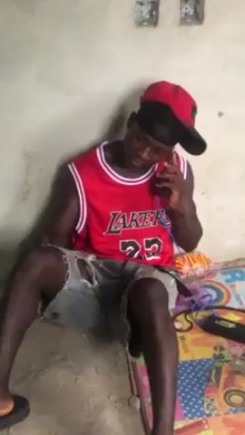A young man wearing a red LA Lakers jersey, ripped denim shorts, and a red and black baseball cap sits on the floor and talks on a cell phone. He is leaning against a white wall. A colorful mat is on the floor beside him. The man appears to be in a room with exposed wiring near the ceiling.