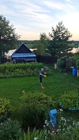 A person wearing a green shirt and dark shorts is visible in a grassy backyard, in what appears to be a rural setting. The person is standing near some plants, and there are several other objects in the yard, including a fence, a shed, and some barrels. The sky is visible in the background, and it appears to be dusk or early evening. The person is in motion, and it is unclear what action they are performing. The video is shaky and out of focus in places.