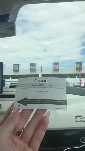 The video shows a close-up of a person's hand holding up a Sanef toll ticket in front of a toll plaza. The hand has long, manicured nails with a pink and white design. The ticket is white with black and gray text and has the Sanef logo at the top. The text on the ticket is in French and reads: "VALIDITE DU TICKET = 24 HEURES" (Ticket validity = 24 hours), "En cas d'absence du ticket ou dépassement du délai de validité du ticket" (In case of absence of the ticket or exceeding the validity period of the ticket), and "TARIF LE PLUS CHER" (Most expensive rate). There's also an arrow pointing left with text that translates to "Any ticket issued at the entry toll booth must be presented at the exit toll booth or to retrieve a ticket at the entry toll booth." At the bottom, it says "TTS V3 - 04/2024".

The background shows a toll plaza with several booths. Each booth has a screen displaying an icon that appears to be a credit card. The sky is cloudy, suggesting daytime. The scene is likely a highway toll plaza in France, given the Sanef branding and French text on the ticket. The person is likely preparing to pay a toll or has just received a ticket.