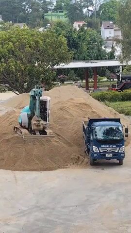 An outdoor scene shows an excavator filling a dump truck with sand. The turquoise excavator stands on a large pile of sand, its arm extended towards a blue dump truck parked adjacent to the pile. The excavator's bucket is dumping sand into the truck bed. In the background, trees, grass, and buildings are visible. A structure with a white roof is partially visible behind the sand pile. A black train car is seen on tracks in the background to the right of the structure. The ground in the foreground appears to be paved.