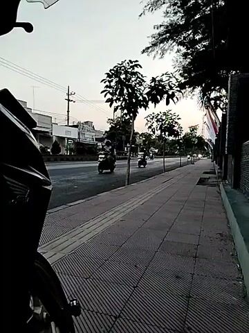 The video shows a street scene with a sidewalk in the foreground. A motorcycle is partially visible in the lower left corner. The sidewalk is paved with rectangular tiles laid in a diagonal pattern, with a textured tactile paving strip running along the edge closest to the road. Several small trees are planted along the sidewalk. Across the street, there are buildings, more trees, and utility poles. Several motorcycles are visible on the road, some with riders. The sky is overcast, suggesting it is either early morning or late afternoon. The overall impression is of a quiet urban street.