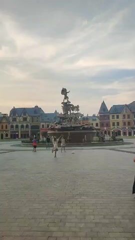 The video shows an open square paved with gray blocks.  In the center of the square is a large, tiered fountain with a sculptural element at its peak. Around the fountain, several people are walking or standing.  The architecture surrounding the square appears European-inspired, with buildings of varying heights and colors featuring gabled roofs and decorative details. People of different ages and clothing styles are present, strolling casually across the square. The sky is overcast with some hints of blue visible.
