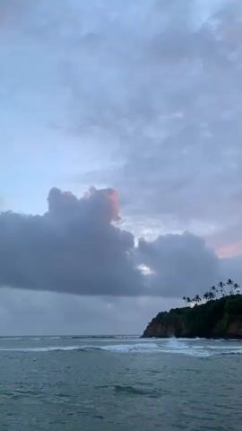 This is a vertical, landscape video of a coastal scene at what appears to be dusk or dawn. The sky is filled with dramatic, textured clouds that are tinged with pink and purple hues, indicating the transition between day and night. A large, dark cloud formation dominates the left side of the frame, while lighter, more diffused clouds stretch across the horizon.

In the middle ground, a lush, green headland juts out into the ocean. The headland is covered in dense vegetation and dotted with numerous palm trees that stand silhouetted against the sky. A few buildings with reddish-brown roofs are visible nestled amongst the trees on the highest point of the headland, suggesting a resort or residential area. The rocky coastline is visible at the base of the headland, with waves crashing against it, creating white foam.

The foreground is dominated by the ocean, which is a deep, textured blue-green. Gentle waves with white crests are rolling towards the shore. The water appears somewhat choppy, reflecting the dynamic nature of the sky.

The overall mood is serene and slightly melancholic, with a sense of natural beauty and tranquility. The combination of the dramatic sky, the verdant headland, and the ocean creates a picturesque tropical setting. The time of day is clearly either sunrise or sunset due to the colored sky and lighting conditions. There are no people or text visible in the video. The location context provided, Batawala, Sri Lanka, aligns with the visual cues of a tropical coastline with palm trees and a headland.