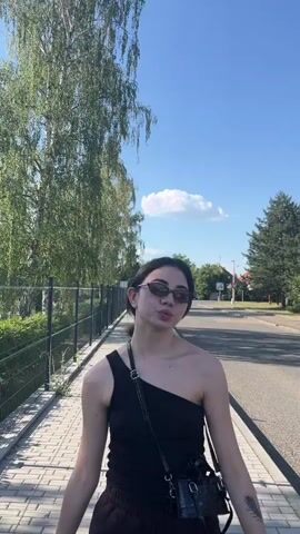 A young woman with dark hair and sunglasses, wearing a black one-shoulder top and dark pants, stands on a paved walkway next to a metal fence. She is holding a small black purse with a strap across her body. The woman smiles and makes peace signs with both hands. Behind her, a paved road stretches into the distance, lined with trees and houses. The sky is blue with a single white cloud. The lighting suggests it is daytime.
