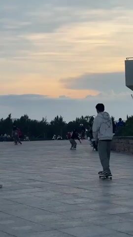 A person on a skateboard moves from right to left across an open plaza with other people in the background. The skateboarder wears a light colored hooded jacket and gray pants. To the left, two small children in front kneel on the ground. The child closer to the camera sits on a skateboard. The children watch the other people around the plaza. To the left, several people walk or run in the open area. Trees are visible in the background. The sky is blue, pink, and white. A light post stands on the right side of the background. A motorcycle is parked to the right.