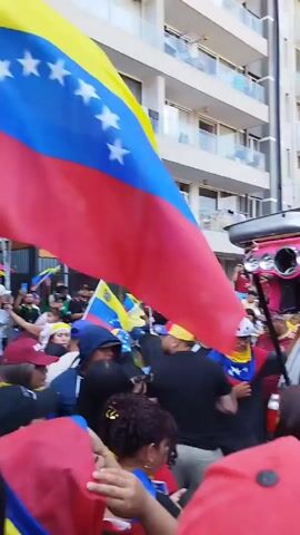 The video shows a large crowd celebrating, many holding the Venezuelan flag and wearing hats or shirts with the same colors.  The open trunk of a car is visible, filled with speakers and adorned with a Venezuelan flag. The people are densely packed in the street, which is lined with tall buildings. The sky is blue, and the atmosphere appears festive. Many power lines are visible in the sky.