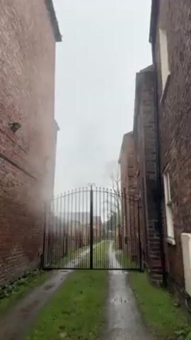 The video shows a view between two brick buildings, looking towards a black metal gate. The ground is wet, with a paved path and patches of grass. The sky is overcast and gray. The brick buildings are tall and narrow, with the gate closing off a road or pathway extending into the distance. There are some trees visible beyond the gate, but they are partially obscured by fog. It appears to be a rainy or damp day.