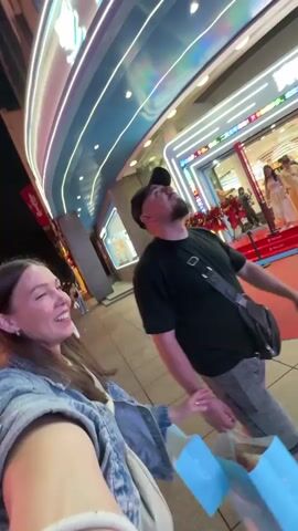 The video shows a woman holding a phone, filming herself and a man walking in front of a store named "Candy Toy". The woman has long brown hair and is wearing a denim jacket. The man has a beard and is wearing a black shirt, grey shorts, and a baseball cap. He is carrying a bag. The store is decorated with red carpets and neon lights. The scene appears to be at night. The camera moves, capturing the surroundings as they walk. The woman smiles at the camera.