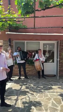 Two musicians play instruments in front of a house. The man on the left wears sunglasses and a white shirt, playing a black accordion. The man on the right wears a white shirt with red embroidery and dark pants, playing a large, light-colored drum. The house has a stone base, an orange upper story, and white windows and doors. A vine-covered trellis is above the musicians, and a decorated staircase leads to an upper level on the right side of the frame.