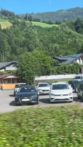 The video shows a view of a village in a mountainous region on a sunny day. In the foreground, there is blurred green foliage. In the middle ground, there is a parking lot with several cars, including a white Mini Cooper, a silver hatchback, and a dark sedan. There is a small orange and brown building with a sign that is difficult to read. Behind the parking lot are several houses, some with solar panels, and lush green trees. In the background, steep, tree-covered mountains rise under a clear blue sky with a faint contrail. A person in a white shirt and shorts is visible standing near the silver car. The camera appears to be moving, possibly from a vehicle.