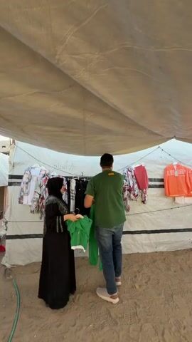The video opens with a close-up of a woman in a black hijab and a man in a green t-shirt holding up clothing under a large, tan tarp. The man is wearing blue jeans and the woman is wearing a black abaya and holding a green t-shirt. The background shows racks of colorful clothing. The camera then pans out to reveal a sandy, open-air space with a tarp covering the top, creating a shaded area. Several people, including children and adults, are visible within this space. In the foreground, two children stand looking towards the camera. To the right, another child in a green shirt stands looking down. In the background, a group of people are gathered, some standing and some sitting. A few more children are visible in the background, some appearing to interact with adults. The scene appears to be some sort of outdoor market or gathering.