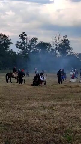 A low-angle, wide shot shows a field with a battle reenactment during dusk. The dry field is light brown, and the sky above is cloudy, with white clouds and some blue sky. On the left side, a person in a white top, brown pants, and a dark cap stands next to a horse that faces left. Another person in a dark top, white pants, and a dark cap crouches nearby, with a long gun. At the center, many people are scattered, some standing and some crouching. A cloud of white smoke fills the air. A person in a dark top and light pants crouches, holding a rifle in front of them. Another person in a white top and dark pants is running towards the right. A person in a red top, white shirt, dark pants, and a red hat stands facing left. Another person in a dark blue top and light pants crouches. Further to the right, a person in a white top and dark pants is kneeling, while another in a white top and dark pants stands with a rifle. More people are in the background. The background also shows trees, and on the horizon, some buildings and cars. The camera pans slowly to the right.