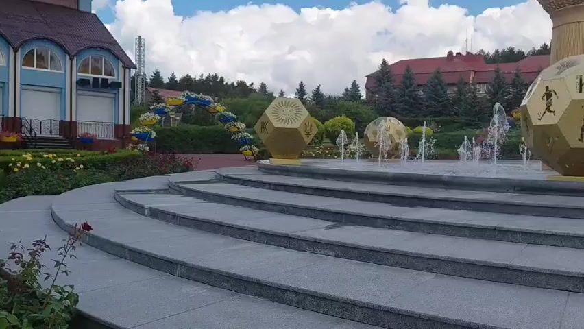 A daytime, eye-level, wide shot shows a fountain with several golden, dodecahedron-shaped structures, some with intricate designs and figures, located in a plaza. The fountain is active, with water jets spraying upwards. Behind the fountain, a large, golden, globe-like structure on a pedestal is visible. The plaza has tiered stone steps in the foreground and is surrounded by trees, buildings with red roofs, and a cloudy blue sky. A curved archway decorated with yellow and blue flowers is visible to the left of the fountain. There are no people in the frame.