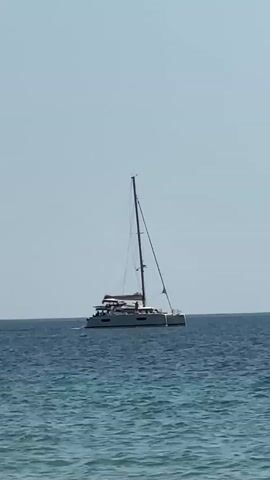 This media file shows a catamaran sailboat on a calm, deep blue sea under a clear, pale blue sky. The boat is white with a tall mast and several sails, one of which is partially unfurled. There are people visible on the deck of the boat, suggesting it is being used for leisure or travel. The water is gently rippled, indicating a peaceful day. The bright sky and clear visibility suggest it is daytime, likely midday, with fair weather. The scene is set on the open water, with no land visible in the frame. The overall mood is serene and tranquil. There is no visible text in this media file.