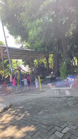 The video shows an outdoor playground area on a sunny day. In the foreground, a brick-patterned pathway is visible. In the background, there is a playground structure with colorful bars and slides. Several children are playing on the playground equipment. Some adults are also present, with one person sitting on a bench and another lying down on what appears to be a mat or blanket. Large trees with lush green foliage are surrounding the playground, providing shade. Sunlight filters through the leaves, creating bright patches and shadows on the ground. The overall atmosphere suggests a relaxed and recreational setting.