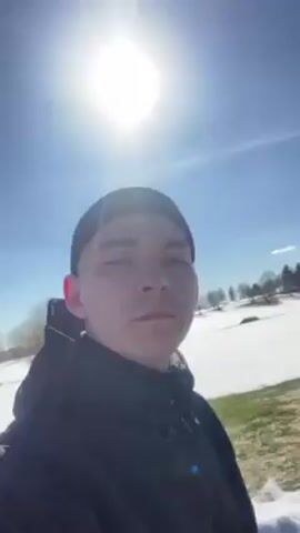 A young man wearing a black beanie and jacket looks at the camera while outside on a sunny day. There is snow on the ground and evergreen trees in the background. The man appears to be walking forward.