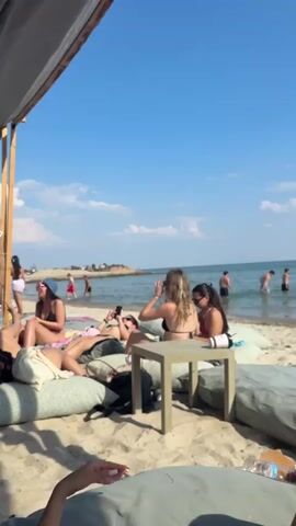 The video shows a sunny day at a beach in Fanari, Greece. The scene is set on a sandy beach with a clear blue sky and calm water. In the foreground, several people are relaxing on large cushions on the sand, some are sunbathing, others are looking at their phones or talking. A small wooden table is placed between them.

In the background, more people are enjoying the water. Some are wading in the sea, while others appear to be swimming or playing in the water. The sea is a gentle blue, and there are a few white clouds scattered across the sky.

The overall atmosphere is one of relaxation and enjoyment on a summer day at the beach. The video captures a typical holiday scene, with people taking advantage of the warm weather and beautiful surroundings. A life preserver ring is visible in the foreground. A wooden post with a sign displaying "1003" is also visible, offering a potential clue to the location. The time of day is clearly daytime, given the bright sunlight and blue sky.
