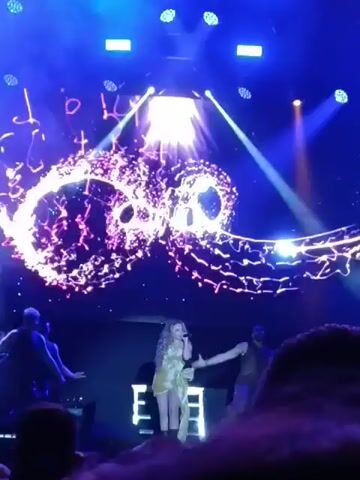 A woman sings on a stage during a performance. The stage is lit with blue and white lights and has a screen behind the singer displaying an abstract swirling design in pink and white. Two dancers are positioned on either side of the singer. The dancers are male and appear to be wearing vests or harnesses, and they are moving their arms. Smoke effects billow around the dancers and singer. In the foreground, a crowd of people can be seen, likely the audience at the performance. The video is shot from within the audience.