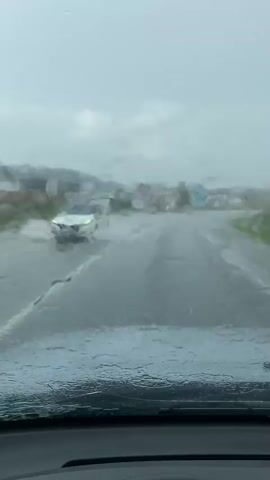 This video shows a view from inside a vehicle driving through flooded roads during a storm. The windshield is wet with rain, and the wipers are intermittently clearing it. The road ahead is covered in water, and houses and trees are visible in the background, some appearing partially submerged. The sky is overcast and gray, indicating heavy rain and poor weather conditions. The vehicle appears to be moving slowly, navigating the flooded conditions.