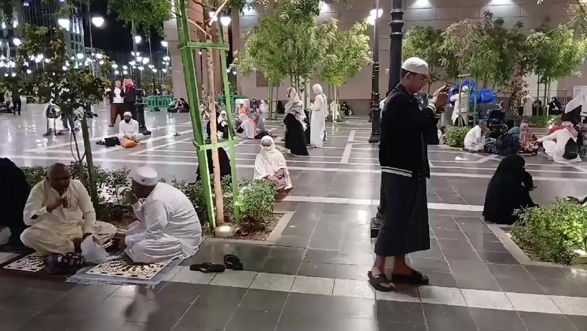 The video shows a tiled plaza at night with many people sitting and milling about. Most of the people are wearing traditional Muslim clothing, including white robes and head coverings for men and black abayas and hijabs for women. Some are sitting on prayer rugs. There are young trees planted in raised beds throughout the plaza, with lights illuminating them. Street lights are visible in the background. One young man in black is standing and looking at his phone. Another man in white walks past the camera.