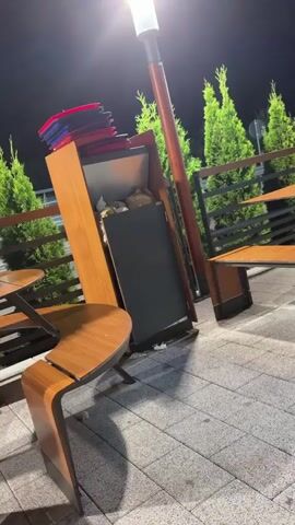 The video shows an outdoor seating area at night. There are wooden tables and benches, some of which are empty, and some have items on them. In the foreground, a wooden table has a pack of cigarettes and some sugar packets. Further back, other tables are visible, with a water bottle on one of them. There are also some bushes and trees in the background. The lighting suggests it is nighttime, with streetlights or building lights illuminating the area.