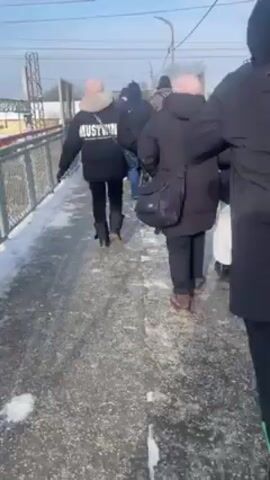 A group of people walk on a snow and ice covered overpass on a sunny day. They are all bundled up in winter coats. Some wear hats and carry bags. The overpass has metal railings. A train and power lines are visible in the background.