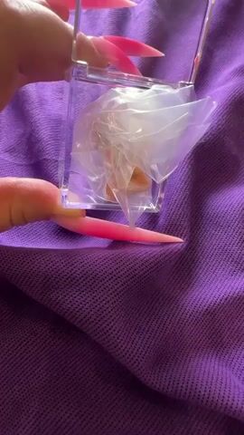 The video shows a close-up of a person's hands with very long, pointed, bright pink and white ombre acrylic nails. The person is holding a small, clear, rectangular plastic box. Inside the box, there appears to be a light brown, possibly powdered substance wrapped in clear plastic. The person is using their long nails to manipulate the plastic wrapping and extract the substance. The background is a textured purple fabric.