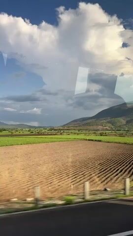 The video depicts a rural landscape from the perspective of a moving vehicle, likely a bus, as indicated by reflections on the window. The scene features expansive fields, some tilled with visible rows of dirt, and others lush green with plants, possibly crops like sugarcane. In the distance, a range of large, rolling hills or mountains with green vegetation is visible. The sky above is partly cloudy, with large white clouds contrasting against a blue sky, particularly on the left, and hints of a brighter, possibly sunnier, sky on the right. A road with a guardrail runs alongside the fields in the foreground, indicating the movement of the camera. The video captures the passage through this scenic countryside.