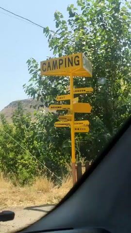 The video shows a yellow signpost with multiple directional arrows. The top arrow says "CAMPING". Below that, other arrows point in various directions with city names like "TIBET", "LONDON", "MOSCOW", "STEPANAKERT", and "MOTEL". The signpost is located outdoors in a rural or semi-rural area with trees and dry grass visible. In the background, there is a hill. The perspective suggests the video is being filmed from inside a vehicle, possibly a car, as part of the windshield frame is visible at the bottom of the screen. The weather appears to be sunny with a clear blue sky. Further down the road, a truck carrying long pipes is seen driving away from the camera. There is also a power pole visible.