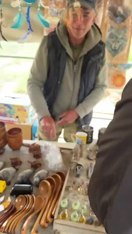 This is a vertical video, likely taken from a mobile phone, showing a close-up of a market stall. The stall is set up outdoors under a canopy, with a backdrop of grassy hills. The foreground is dominated by a wooden table laden with various items for sale.

On the table, there are an assortment of handcrafted goods. These include numerous small wooden bowls and cups stacked together, several plush toys in the shapes of animals like seals and cats, and rows of wooden pens with decorative tips resembling small animals or natural elements. There are also several small, dark-colored carved objects, possibly animal figurines or trinkets, and some keychains. In the center of the table, there is a plastic bag filled with white material. Towards the back, a cardboard box with an image of bananas and the brand name "Terraversa" is visible, along with other smaller boxes.

A man, appearing to be the vendor, is positioned behind the table. He is wearing a dark vest over a green hooded jacket and camouflage pants. He has light-colored hair and is looking towards the camera. In the first part of the video, he is holding a small wooden carved object, possibly a frog, in his left hand and gesturing with his right hand. In the second part, his hands are open, palms up, as he speaks.

Hanging from the canopy above the stall are various decorative items, including dreamcatchers with feathers and beads, and some wall hangings featuring animal images like bears and owls. The overall impression is that of a small, open-air market specializing in souvenirs and handmade crafts. The lighting suggests it is daytime.