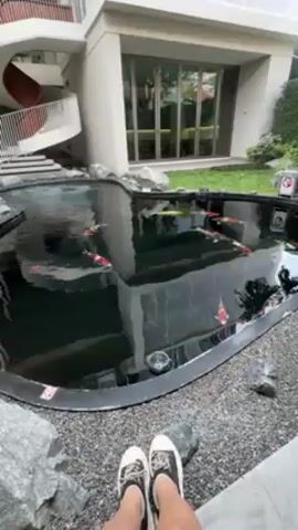 From a low-angle, stationary viewpoint, the video shows a kidney-shaped koi pond set within a landscaped area of a building. The pond's edge is lined with gray stones. Inside the pond, numerous koi fish of various colors, predominantly red and white, swim leisurely. The water is dark, reflecting the building and sky. The surrounding area features a modern building with a staircase, a red slide, and glass doors. In the foreground, a person's legs and feet, clad in black and white sneakers, are visible, suggesting the person is sitting at the edge of the pond.