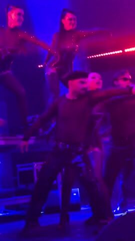 This video captures a live performance on a stage bathed in vibrant blue and pink lighting. The stage is set up with what appear to be elevated platforms on the left side, occupied by two female performers in shiny, form-fitting black outfits with cutouts and thigh-high stockings. They are striking dynamic dance poses. In the foreground, a male performer, wearing a black mesh shirt, black pants, and a black beret, is in the center, also dancing energetically. To his right, another male performer in sunglasses and a similar black mesh shirt and pants is also dancing, and to the far right, another individual in a black outfit is partially visible. In the background, a large screen displays a fragmented, broken-glass-like visual effect. The overall atmosphere suggests a high-energy musical performance.