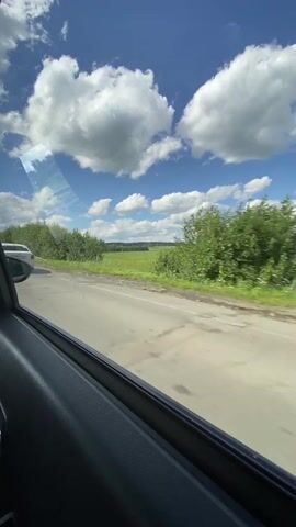 This video shows a view from inside a vehicle traveling on a road. The camera is positioned to look out the side window, capturing the passing scenery. A white sedan is seen in the distance in the first few seconds, followed by the back of a large green trailer truck. The road is paved, and there is a guardrail on the right side of the road. Beyond the guardrail, there is a lush green field with trees in the background. The sky is bright blue with fluffy white clouds, indicating a sunny day. The interior of the vehicle, including part of the door panel and window frame, is visible in the foreground. The overall impression is a scenic drive through a rural landscape on a clear day.