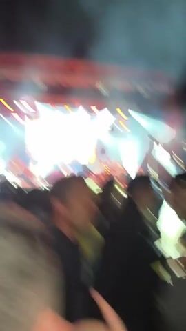 A crowd of people watches a live band performing on a stage. Bright white lights shine on the band. Above them, a large screen shows images, including one of Bob Marley. The band is playing instruments, and the audience has their hands up in the air. The environment appears to be a large indoor venue.