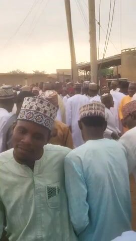 The video displays a crowded outdoor scene in what appears to be a town or city, consistent with the provided context of Jogana, Nigeria. Numerous people, predominantly men, are gathered closely together in a street or open area. Many are dressed in traditional attire, including various styles of kufis or caps, which are often embroidered or come in solid colors like white and red. Some wear long, flowing garments, while others are in shirts of light green, light blue, brown, and white.

The background features several buildings, some with white exteriors, suggesting a residential or commercial district. Overhead, utility poles and power lines are visible, along with tall streetlights. In one segment (0:03-0:04), a yellow three-wheeled vehicle, resembling an auto-rickshaw or tricycle, is partially visible among the crowd, and mats or sacks are laid on the ground.

The activity shows people standing, mingling, and moving slowly through the dense gathering. Several individuals are seen conversing, and at least one person is holding up a smartphone, possibly to record the event. The lighting suggests it is late afternoon or early evening, with a bright, hazy sky casting a soft, diffused light over the scene. The overall atmosphere appears calm and social, indicative of a community gathering or a busy public space. There is no discernible text visible within the video.