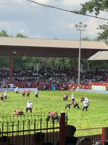 A high-angle, medium shot captures a wrestling event taking place on a grassy field. Numerous athletes, dressed in traditional wrestling attire, are engaged in matches across the field. Spectators fill the tiered seating area under a large, open-air structure. The environment appears to be an outdoor stadium with a bright, cloudy sky. Several individuals in white shirts are present, possibly officials or trainers. The overall atmosphere suggests an active sporting competition.