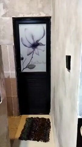 The video shows a close-up, shaky view of a dark-colored door with a frosted glass panel featuring a large, ethereal flower design. The text "Digital Dream Utopia" is visible within the flower design. A small, dark-colored rug lies on the floor directly in front of the door. The walls surrounding the door appear to be rough and unfinished, with some tiling visible to the left of the door. There is a small, dark rectangular object mounted on the wall to the right of the door. The lighting is dim, and the overall impression is of an interior hallway or entryway. No people or explicit actions are captured.