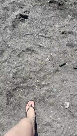 A first-person perspective video shows a person walking on a dark, sandy beach. The camera is pointed down at the person's feet, one of which is wearing a flip-flop. Footprints are visible in the sand, indicating the person is walking forward. The sand appears to be coarse and contains small pebbles. The lighting suggests it is daytime.