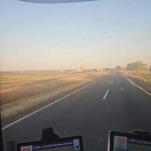 The view from a vehicle traveling on a highway shows the roadside with tall dry grass and a railway track. A large, yellow, figurative sculpture that resembles a person carrying briefcases walks alongside the highway. Several trucks are parked near the sculpture. The sky is light blue.