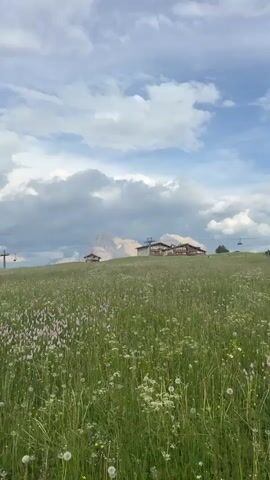 This vertical, eye-level, medium shot captures a serene alpine meadow in Kastelruth, Italy. The foreground is dominated by a vibrant expanse of tall green grass interspersed with delicate white and pink wildflowers, some of which appear to be dandelions in their fluffy seed stage. A few small yellow flowers are also visible amongst the greenery.

In the mid-ground, a gently sloping hillside rises towards a cluster of buildings. The largest structure is a substantial hotel or lodge with a light-colored facade and a dark roof, featuring multiple windows and balconies. To its left, a smaller, rustic wooden cabin with a red roof sits nestled on the slope. The presence of ski lift infrastructure, including pylons and cables, suggests the location is a popular destination for both summer and winter activities, though the lush green landscape indicates it is likely late spring or summer. The vast, open sky above is filled with dramatic, rolling cumulus clouds, some dark and heavy, hinting at the possibility of rain, while patches of bright blue sky peek through, suggesting a dynamic weather pattern. In the distance, a majestic mountain range, possibly the Dolomites, rises majestically, its peaks partially obscured by the clouds, lending a sense of grandeur to the scene.

The overall atmosphere is one of peaceful natural beauty and tranquility, with a touch of remoteness. The lighting suggests it is daytime, likely mid-morning or afternoon, given the brightness of the sky and the definition of the landscape. No people are visible, emphasizing the unspoiled, natural aspect of the location. There is no visible text in the media file.