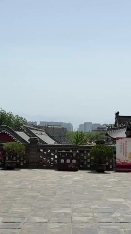The camera pans across a stone courtyard in what appears to be a Chinese historical site. In the background, there is a hazy view of a city skyline. The courtyard features traditional Chinese architecture with tiled roofs, brick walls, and decorative elements. There are potted plants scattered around the courtyard. Two promotional banners with Chinese characters and images of pagodas stand near a trash can. Further along, there is a covered seating area where two men are seated at a bench under a beige canopy. Next to the seating area is a small, booth-like structure selling souvenirs. A man in a black shirt and shorts walks past the booth. The sky is clear and blue, suggesting it is daytime.