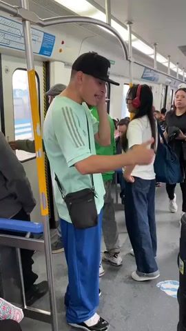 The video captures an interior view of a train or subway car filled with passengers. In the foreground, a young man wearing a light green t-shirt with Adidas stripes, blue pants, a black baseball cap, and a black crossbody bag is talking and gesturing towards a young woman. She is wearing a white t-shirt, loose blue jeans, and red over-ear headphones, and is facing him.

To their right, a large black and orange portable speaker is placed on the floor, emitting loud music. A man in a blue polo shirt and black pants stands nearby, holding a mobile phone and looking towards the speaker. Further back, another man in a grey t-shirt and plaid pants, wearing a reversed baseball cap, is also observing the scene.

The train car is moderately crowded, with several other individuals standing and sitting in the background. Some are looking at their phones, while others appear to be observing the interaction around the speaker. The floor has blue and white markings. The windows show a blurry exterior, indicating the train is in motion. The overhead lighting is bright, illuminating the interior. The overall atmosphere suggests a casual public transport setting, with the music from the portable speaker being a prominent feature of the scene.