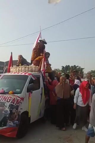 The video shows a white truck decorated with Indonesian flags and streamers, carrying people in traditional attire and what appears to be a large display of produce, possibly fruits or vegetables. A crowd of people is gathered around the truck, with some reaching out towards the truck and others cheering. One person on top of the truck, wearing a conical hat, is interacting with the crowd. The scene appears to be a festive or celebratory event. The sky is clear and bright, suggesting daytime.