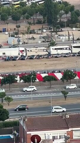 The video is an aerial view of an outdoor event. In the foreground, there is a building with a red tiled roof. Further back, a road runs horizontally across the frame with several white cars driving on it. Behind the road, there is a red and white striped banner with people standing behind it. Beyond the banner, there are several RVs and buses parked. In the background, there are trees and more vehicles. The lighting suggests it is daytime, possibly late afternoon or early evening.
